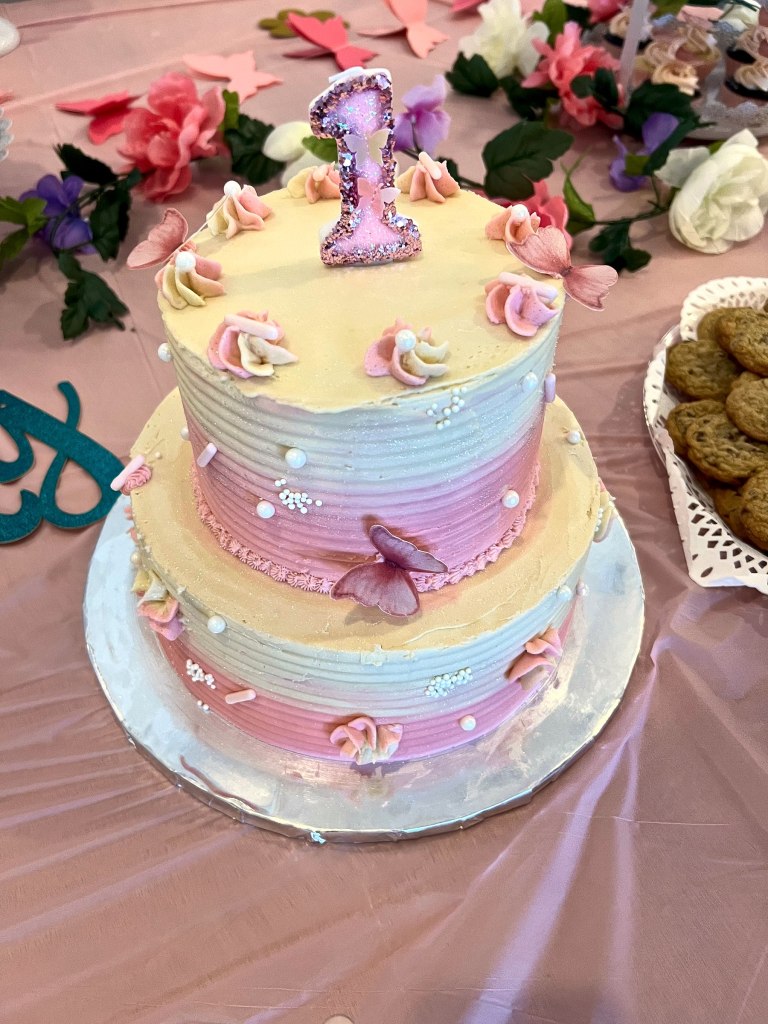 Two-tier pastel first birthday cake with butterflies, frosting rosettes and a sparkly number 1