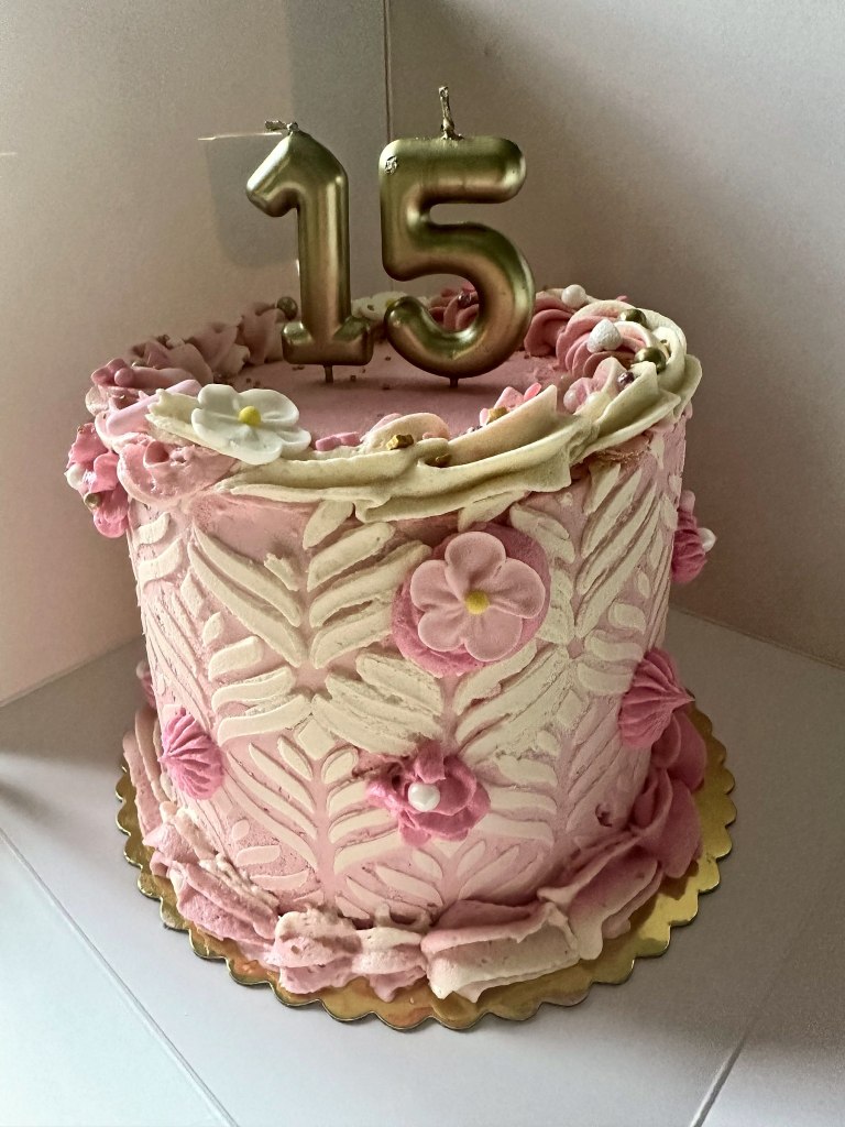 Tall pink and white cake with a textured leaf pattern, sugar flowers and gold number 15 candles