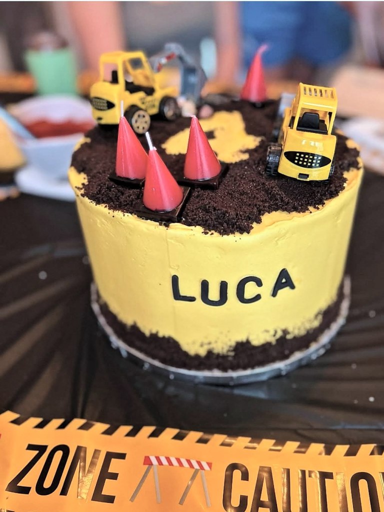 Yellow construction cake with a cookie crumb dirt top, toy diggers, frosting cones and a name in black letters