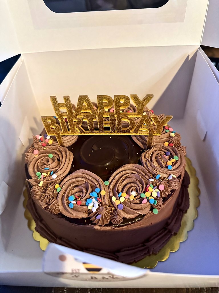 Chocolate birthday cake with chocolate rosettes, rainbow sprinkles and a gold Happy Birthday topper