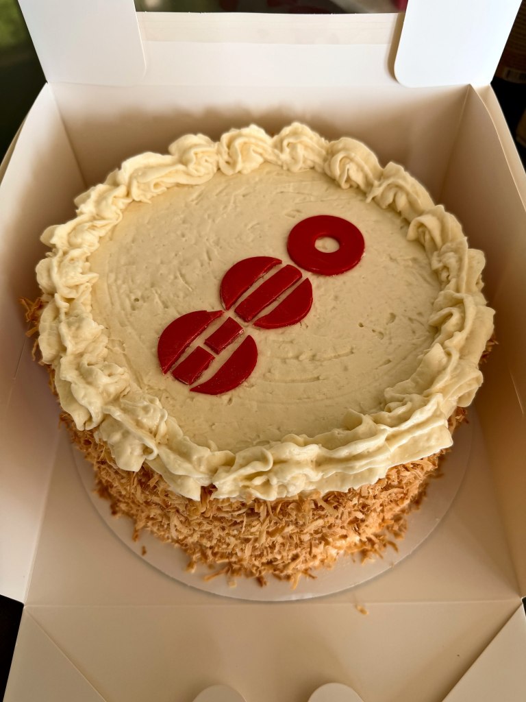 Cream frosted cake with a red number 80 on top and toasted coconut on the sides