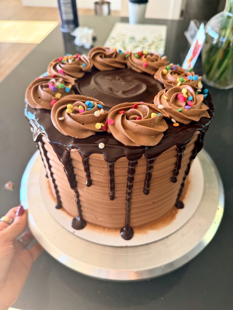 Tall chocolate cake with a chocolate drip and frosting rosettes topped with rainbow sprinkles