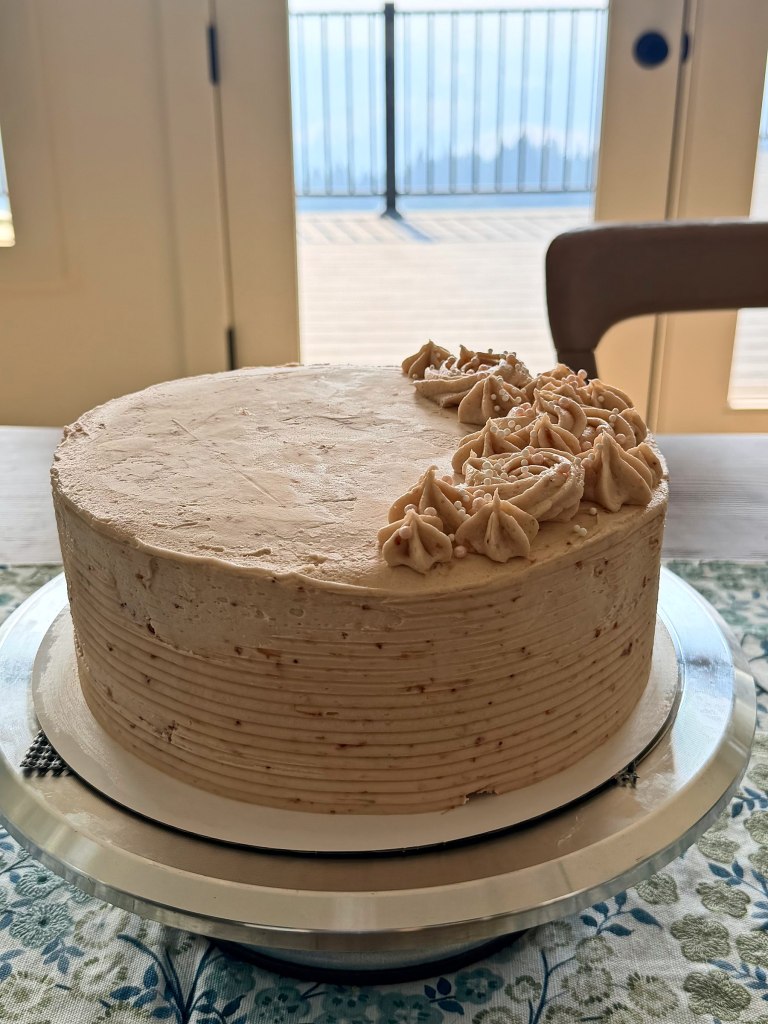 Round cake in light brown frosting with rosettes on one side, by a sunny window