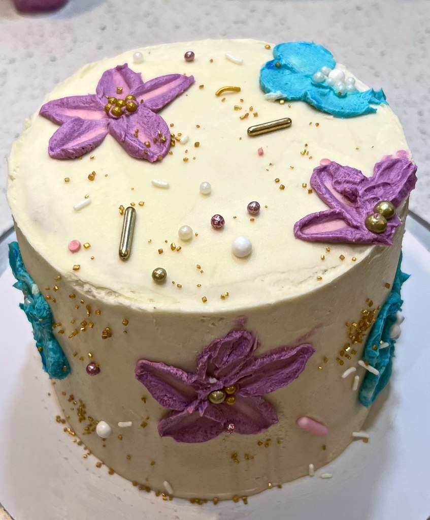 White cake with purple and blue frosting flowers, pearls and gold sprinkles