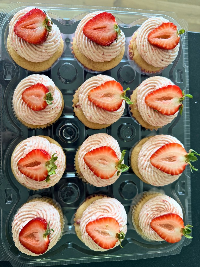A dozen cupcakes with swirled pink frosting, each topped with half a fresh strawberry