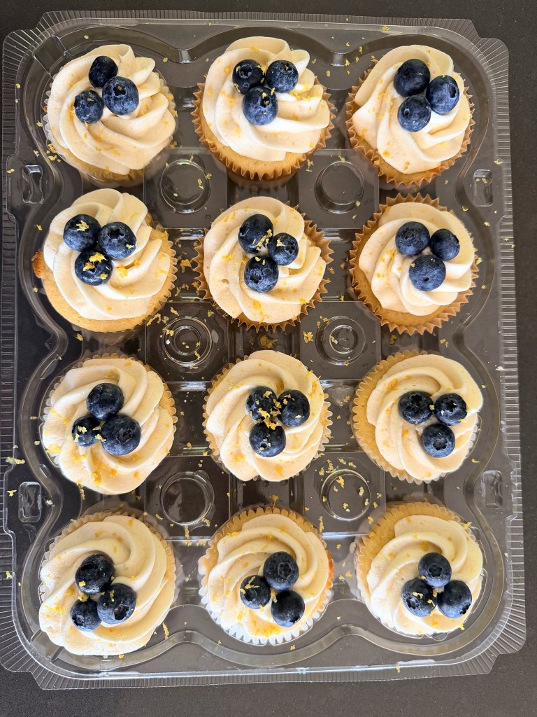 A dozen cupcakes with cream frosting, fresh blueberries and lemon zest